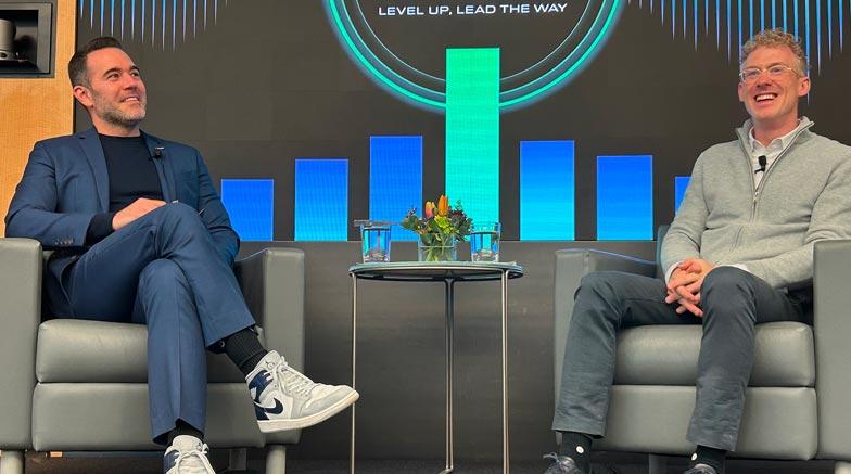 Two men sitting on stage during a panel. Level up. Lead the way is on the screen in the background.