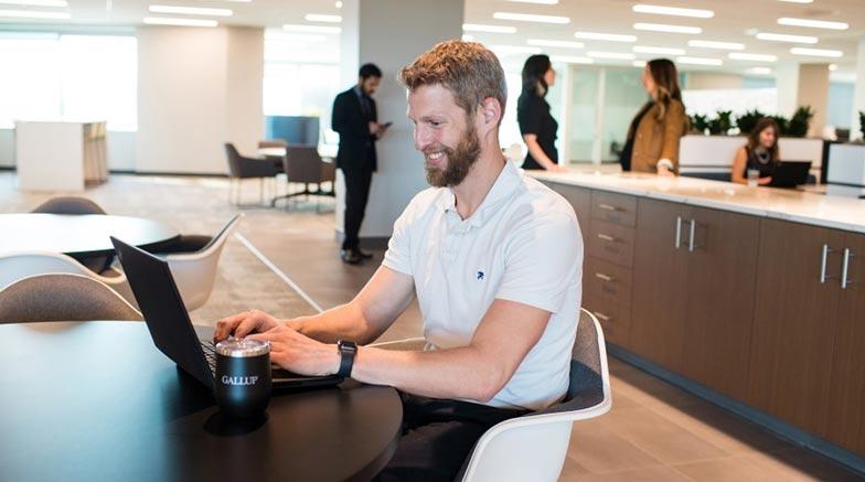 A male sitting at a table working on a laptop with other employees in the background.