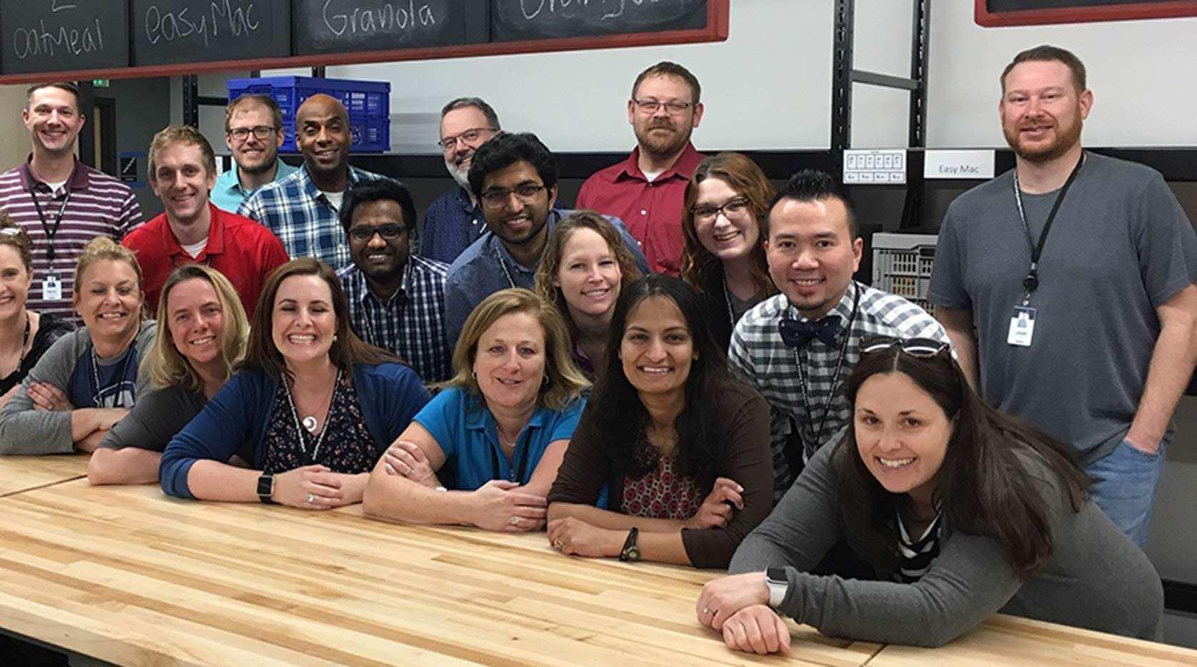 A large group of employees standing behind a long set of tables with chalk boards above and behind them.