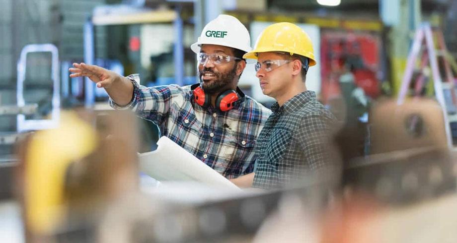 Two industrial workers wearing hard hats and safety glasses review blueprints in a manufacturing facility, with one pointing to direct the other's attention.